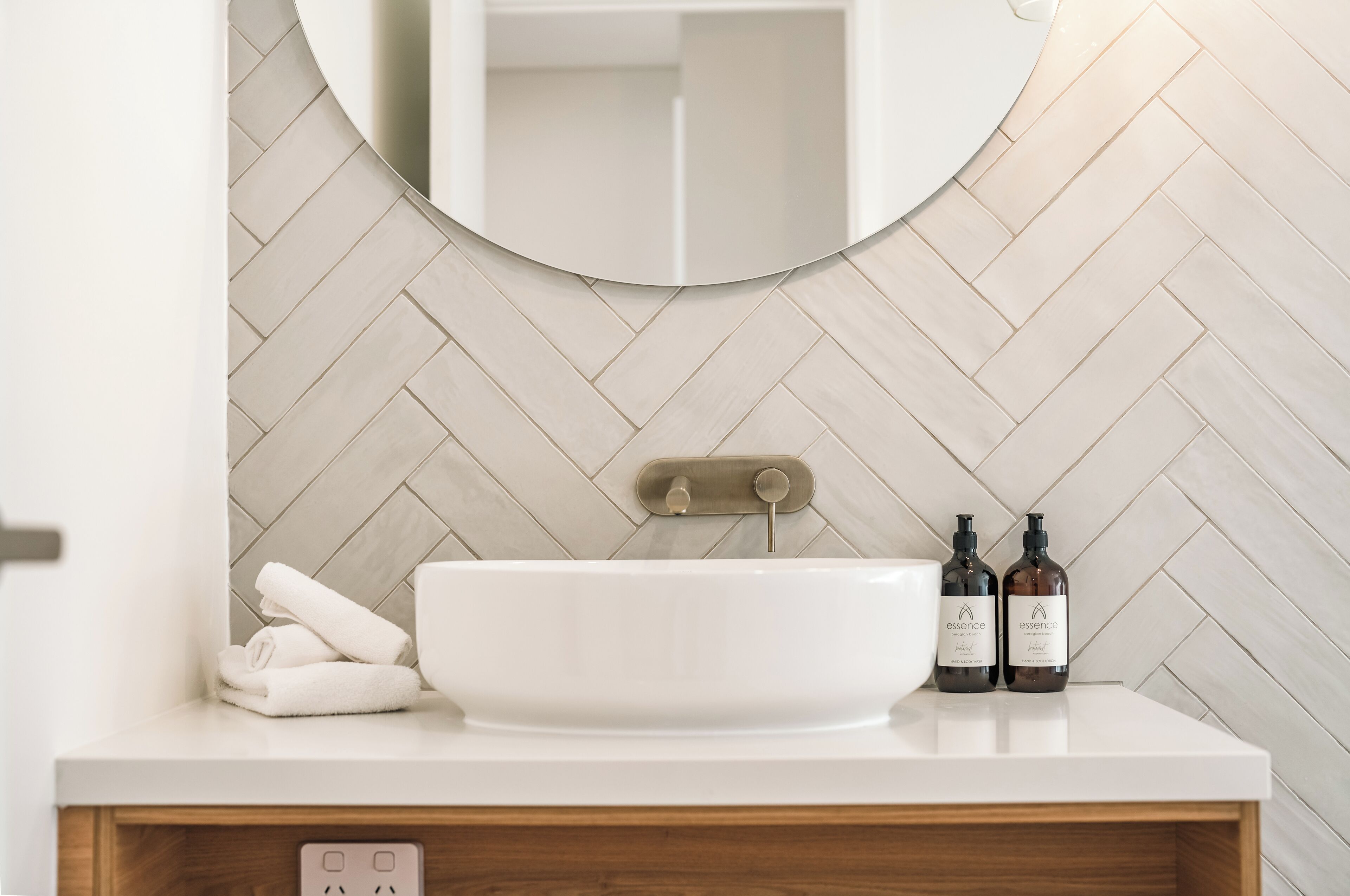 Une salle de bain avec un lavabo blanc, deux bouteilles de gel douche Essence et une serviette posée sur le comptoir.