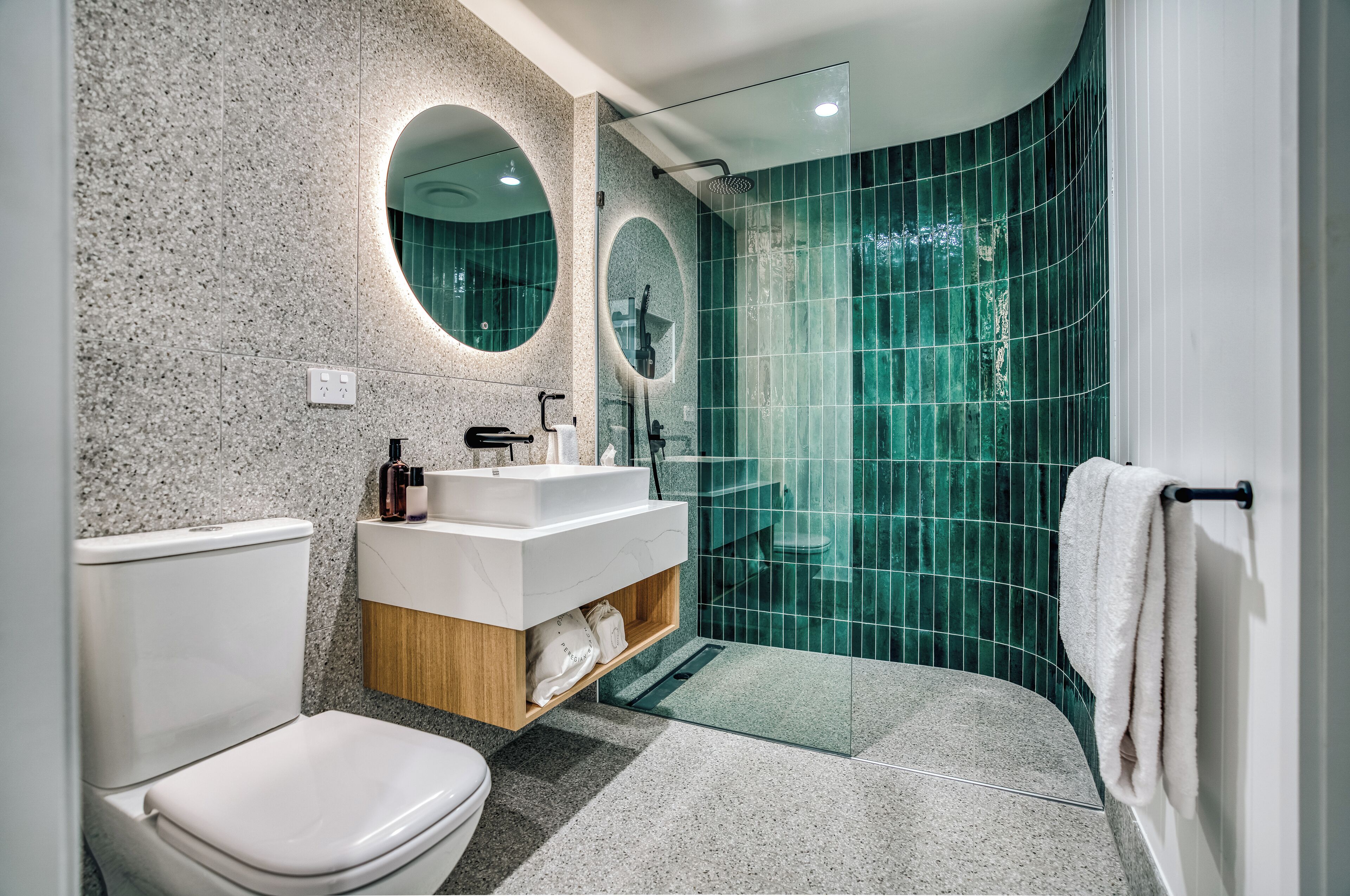 A modern bathroom with a toilet, a vanity with a mirror, and a shower with green tiles.