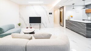 Un salon moderne doté d’un téléviseur à écran plat, d’un canapé, d’une table basse et d’un coin cuisine avec un plan de travail en marbre et des luminaires suspendus.