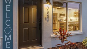 A house entrance with a dark brown door, a 'WELCOME' sign, a window with a view of a kitchen, and a small plant in the foreground. - Bahama Breeze | Private Pool w/ Patio + Outdoor TV! (Sarasota)
