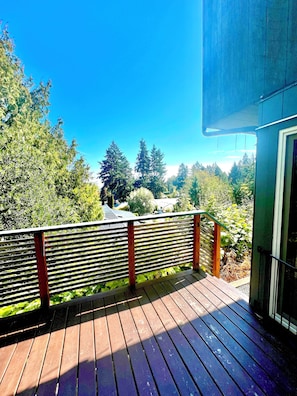 A wooden deck with a metal railing, overlooking a forested area with tall trees and a clear blue sky. - Cozy Cordova Bay Suite - 5min from beach! (Victoria)