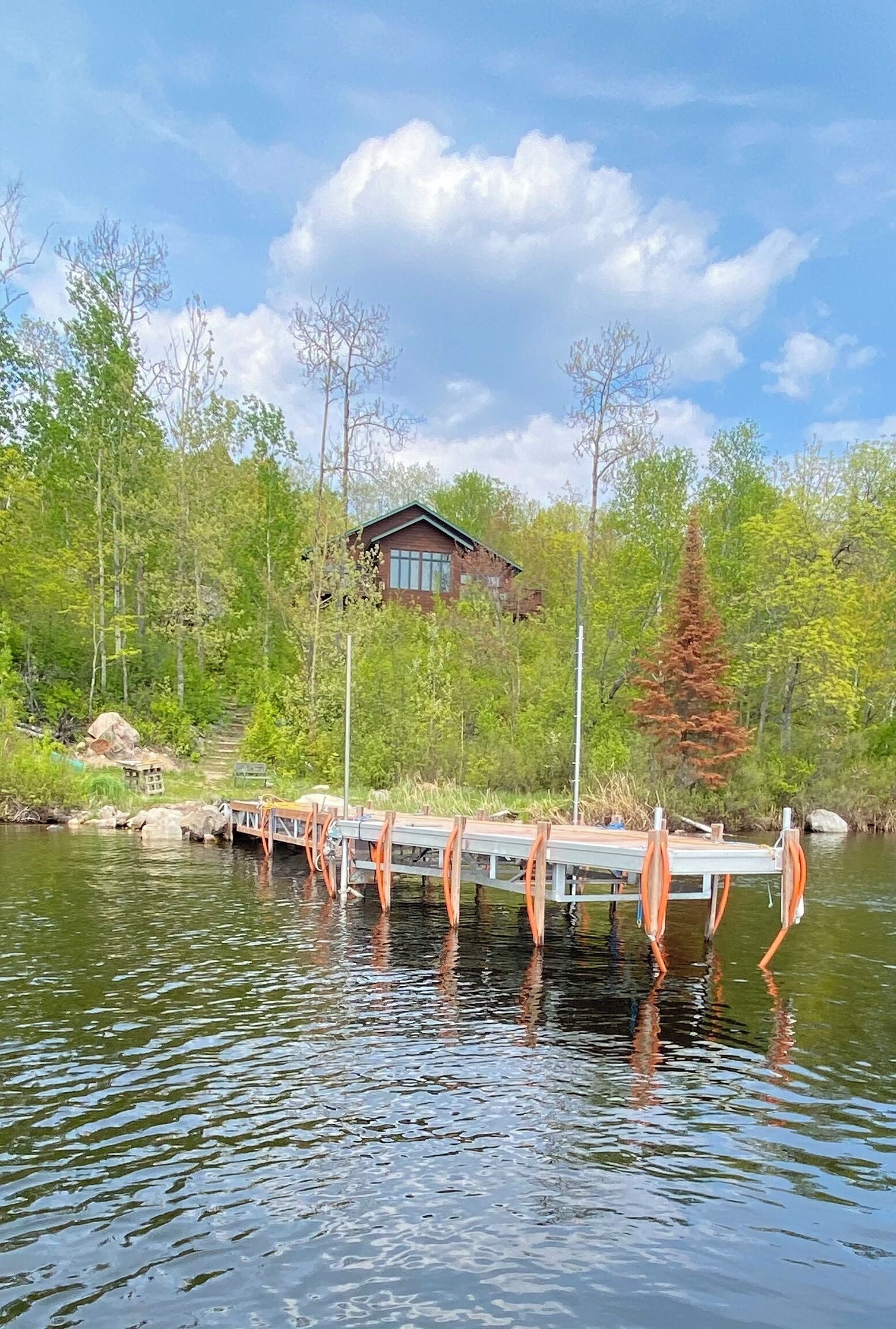 A wooden dock extending into a calm lake, surrounded by lush greenery and a cabin in the background.