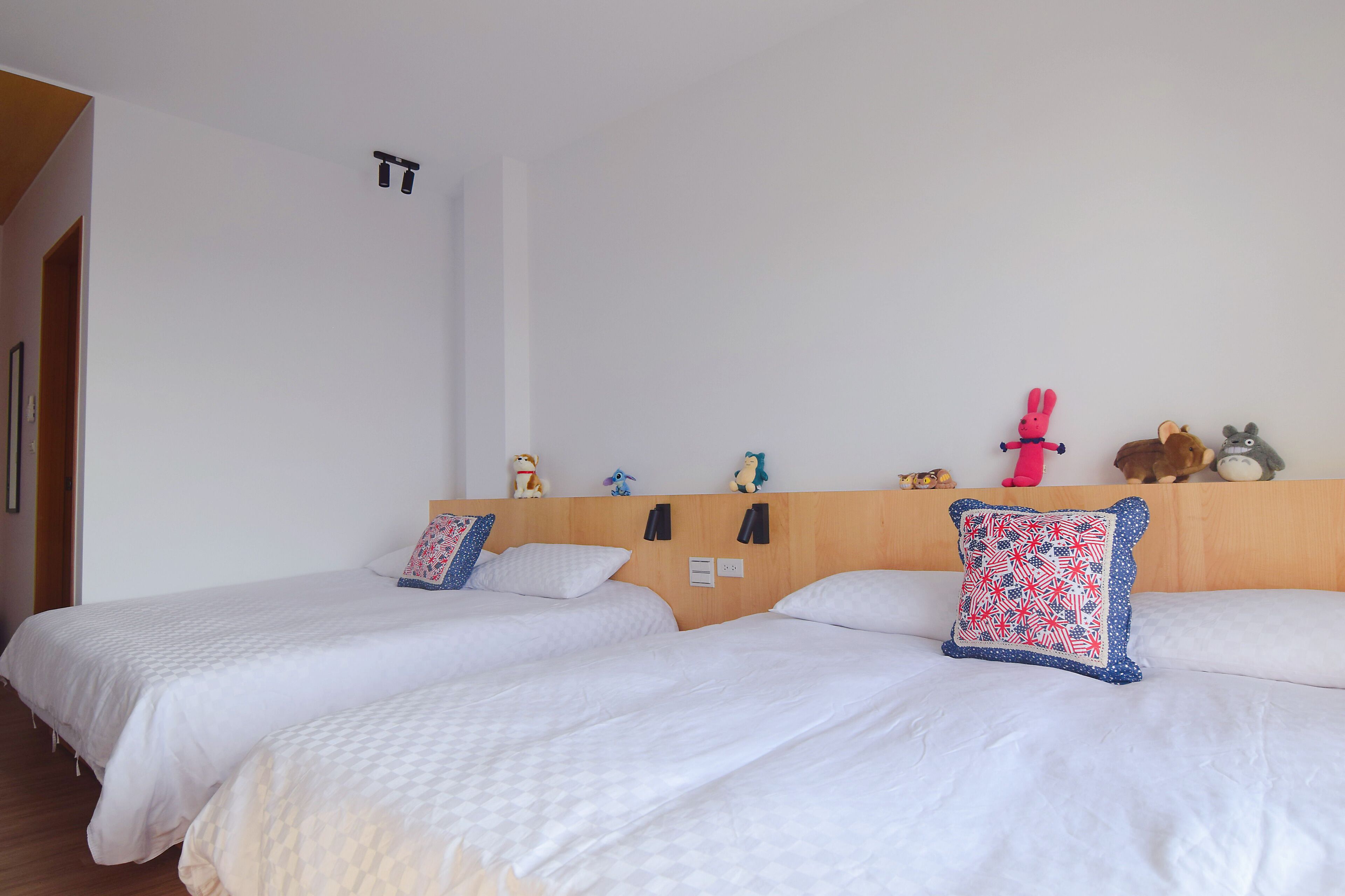 A hotel room with two beds, each with white bedding and decorative pillows. There are wall-mounted light switches and a wooden headboard.