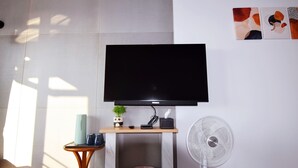 Uma sala de estar moderna com uma televisão de tela plana na parede, uma pequena mesa de madeira com uma planta, um ventilador de chão branco e uma lixeira.