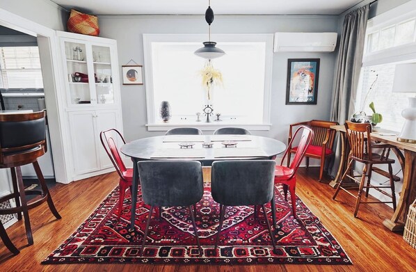 Un coin repas avec une table ronde, des chaises rouges et grises, un tapis à motifs et une fenêtre agrémentée d’un luminaire suspendu.