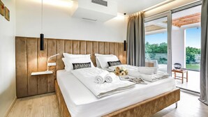 Un dormitorio moderno con cabecera de madera, una cama grande con ropa blanca, una mesita de noche con lámpara y vistas al exterior a través de una puerta corredera.