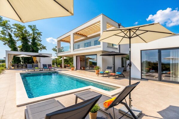 A modern two-story house with a swimming pool, outdoor seating, and large windows.