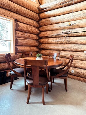 Interno di una baita di legno con un tavolo da pranzo in legno circondato da sedie, una piccola pianta sul tavolo e una finestra con persiane.