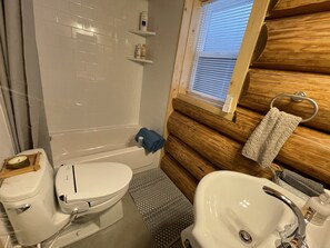 Un bagno con il water, un lavandino e una doccia. È presente un portascopevoli con un asciugamano, una finestra con persiane e uno scaffale con articoli da toilette.
