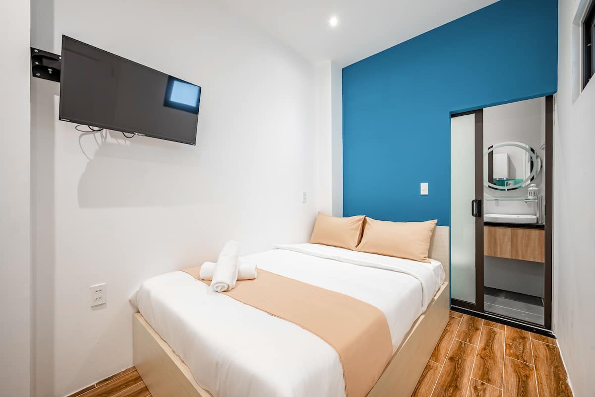 A neatly made bed with a beige and white bedspread, a flat-screen TV mounted on the wall, and a bathroom with a mirror and sink.