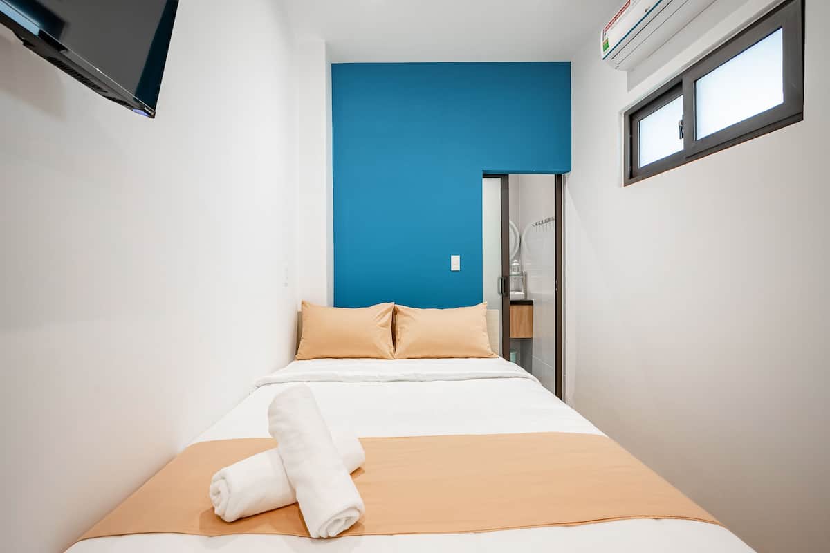 A small, modern hotel room with a bed, a blue accent wall, a window, and a door.