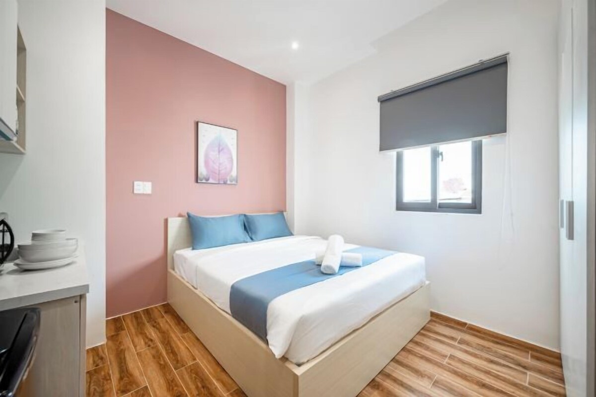 A bedroom with a bed, a window with blinds, a picture on the wall, and a small table with plates.
