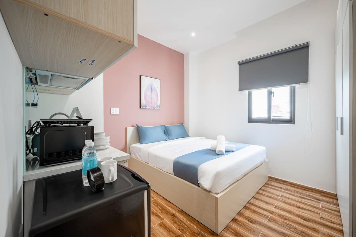 A compact hotel room with a bed, a desk with a computer, a small kitchenette, and a window with a blind.