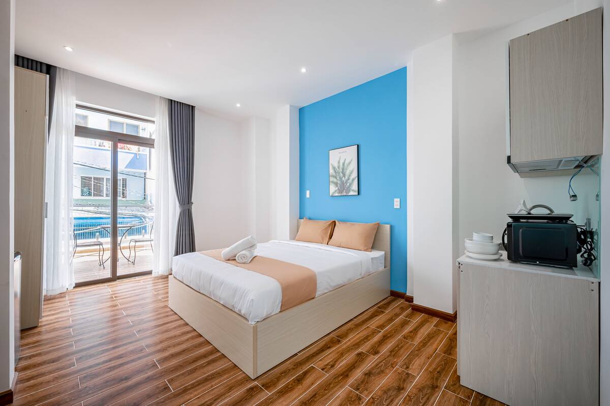A hotel room with a bed, a kitchenette, and a view of a balcony.