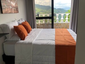 A hotel room with a bed, a painting on the wall, and a balcony with a view of green hills. - Cozy 2-BR Apt with mountain view (Antioquia)