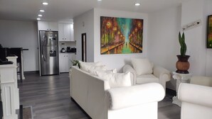 Una sala moderna con un sofá blanco, una zona de comedor con mesa y sillas, y una cocina con refrigerador y armarios.