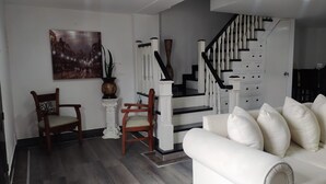 Una sala de estar con un sofá blanco, dos sillas de madera, una escalera con barandillas blancas y un cuadro de un paisaje urbano en la pared.