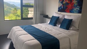 Un dormitorio con una cama grande, vistas a zonas verdes y edificios, un cuadro en la pared y una ventana con cortinas.