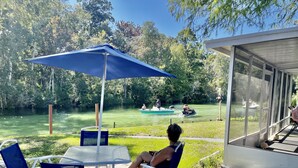 A patio area with a table and chairs, a blue umbrella, and a view of a river with boats. - Spring 12% Discount-April May: The Hidden Jim River Cottage -4 Kayaks (Spring Hill)