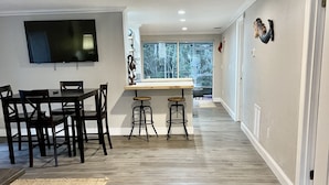 A modern living room with a dining area, a flat-screen TV mounted on the wall, and a large window providing a view of the outdoors. - Spring 12% Discount-April May: The Hidden Jim River Cottage -4 Kayaks (Spring Hill)