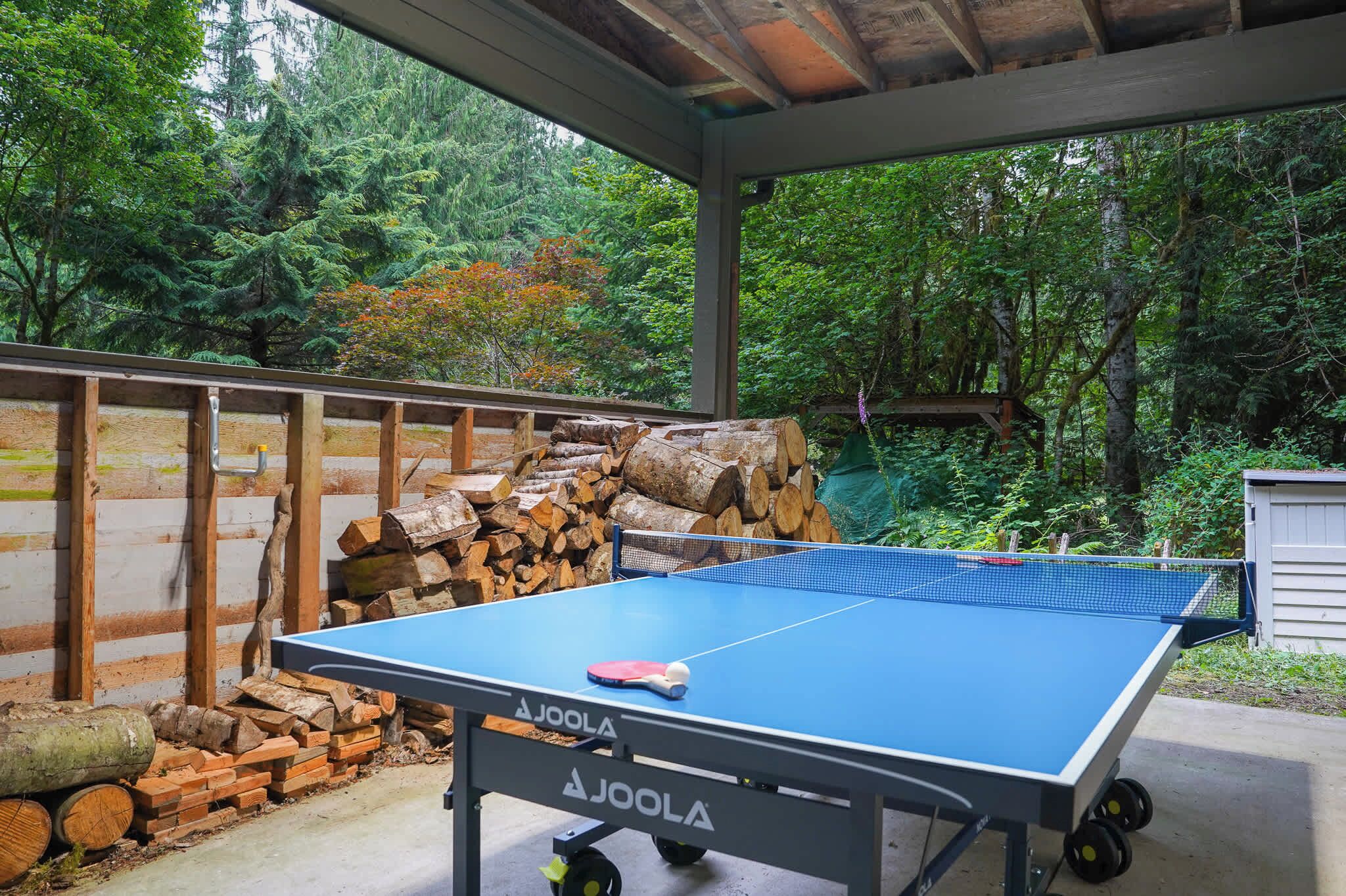 An outdoor ping pong table with a net, surrounded by stacked firewood and a wooden fence.