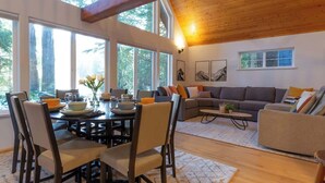 A spacious living area with a dining table, chairs, a sofa, and a coffee table. Large windows provide natural light and views of the outdoors.