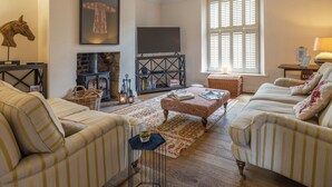 A cozy living room with striped sofas, a fireplace, a television, and a large window with shutters. - Westhill House (Morpeth)