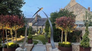 A garden pathway leading to a stone building with a traditional roof, surrounded by potted plants and a unique metal sculpture. - Westhill House (Morpeth)