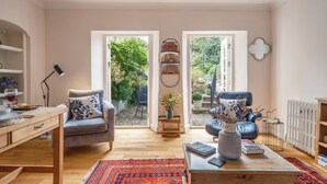 A cozy living room with a sofa, armchair, and coffee table, a bookshelf, and a view of a garden through large windows. - Westhill House (Morpeth)