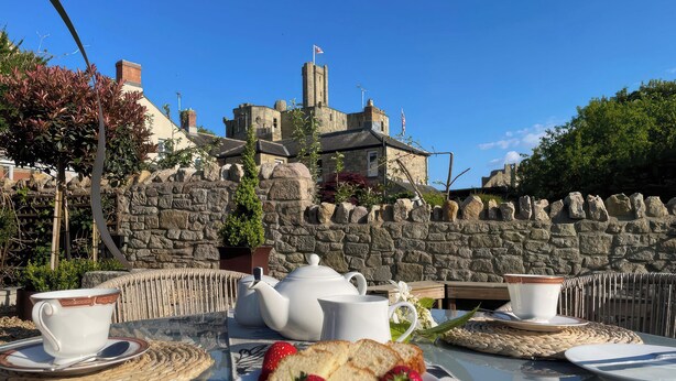 An outdoor dining area with a stone wall, a view of a historic building, and a table set with a teapot, cups, and strawberries. - Westhill House (Morpeth)