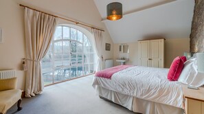 A bedroom with a large window, a bed with white bedding and a pink throw, a bedside table with a lamp, and a wooden headboard.