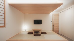 Habitación minimalista con suelo de esteras de tatami, una mesa de madera baja con cojines, televisor montado y puerta corredera.
