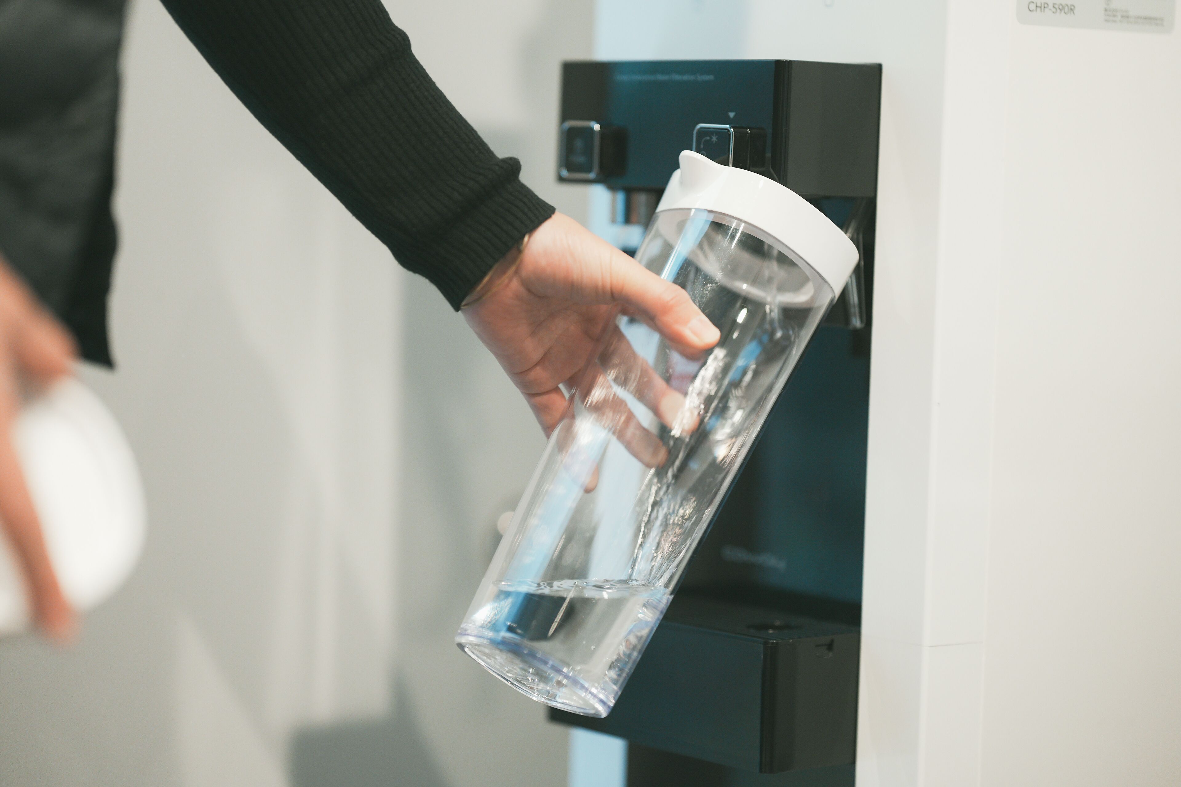 Una mano sostiene una botella de agua transparente con tapa blanca, frente a un dispensador de agua.