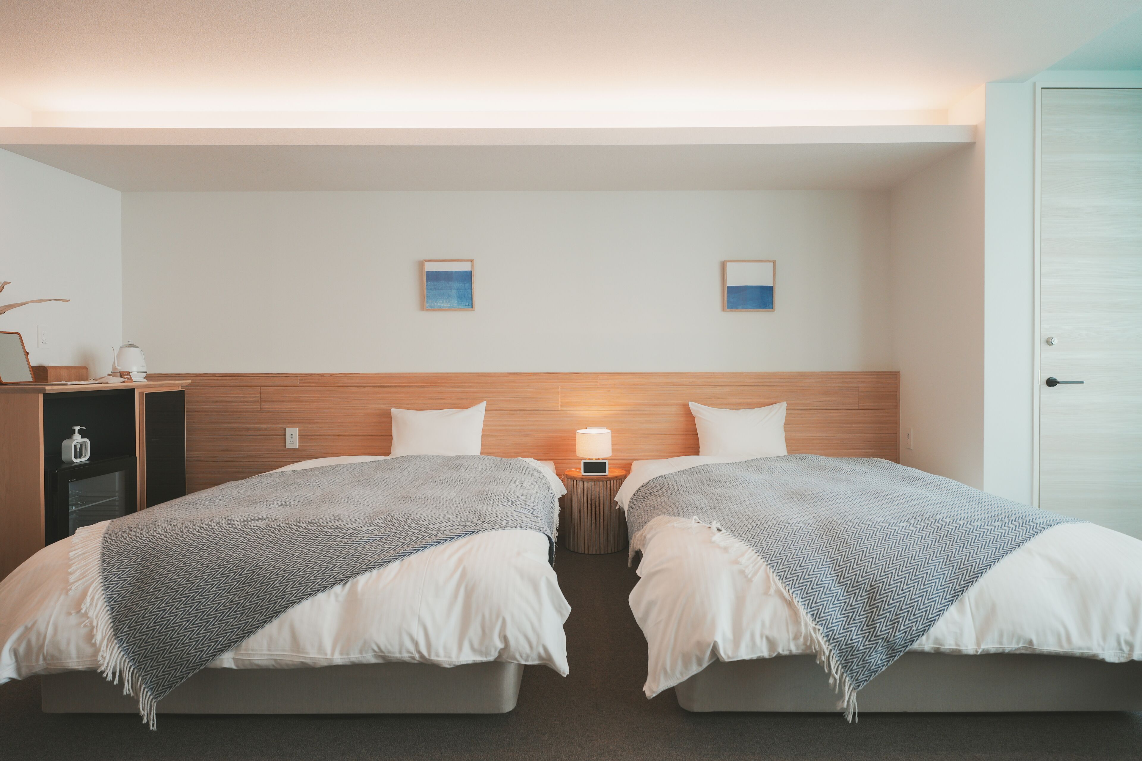 A hotel room with two beds, a wooden headboard, a nightstand with a lamp, and a small shelf with toiletries.
