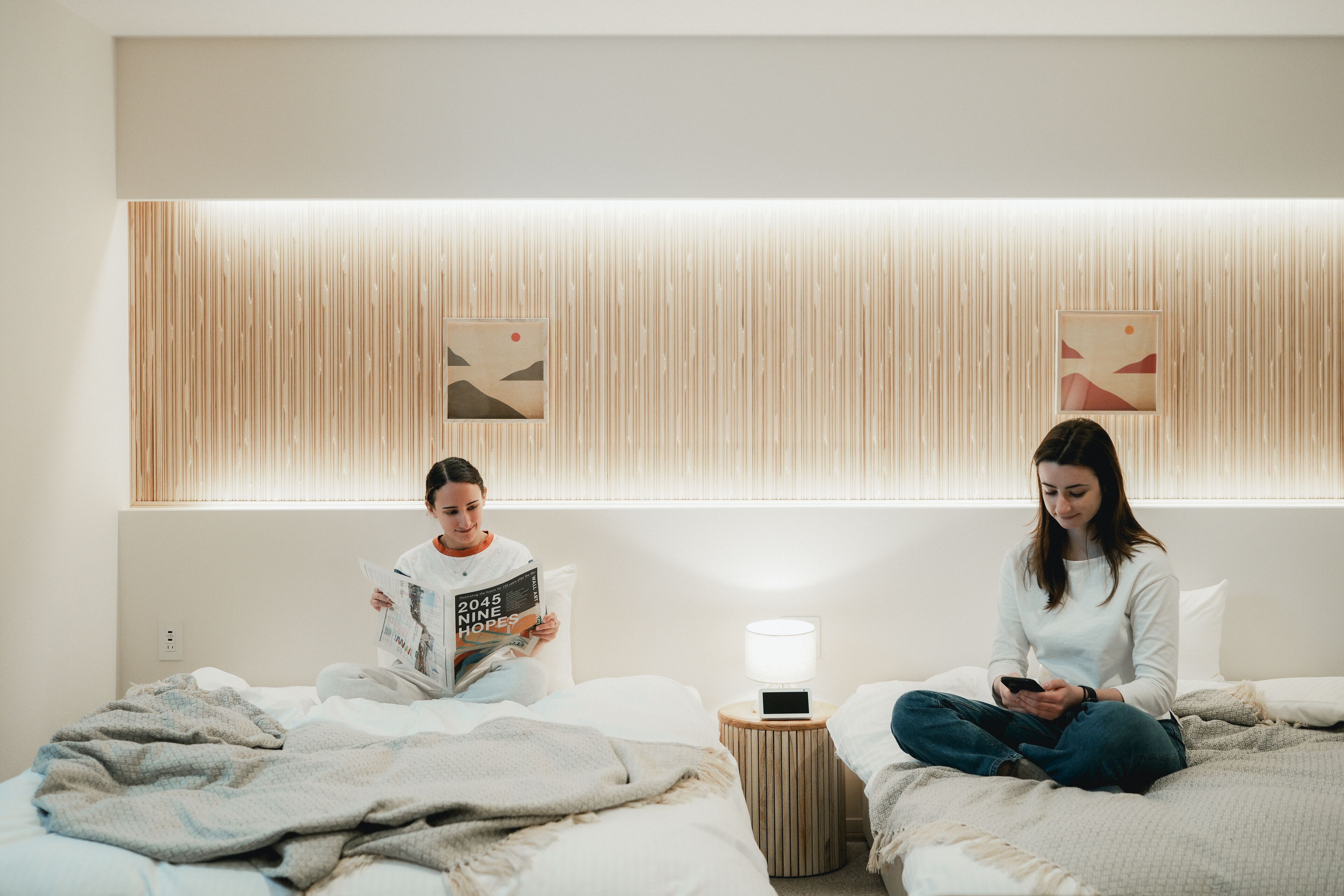 Two people sitting on separate beds in a room with a wooden slat wall, a bedside table with a lamp, and a power outlet.