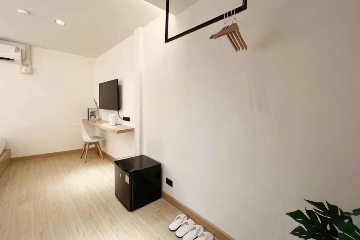 A modern hotel room with a wooden floor, a black refrigerator, a desk with a chair, a flat-screen TV mounted on the wall, and a plant.