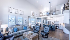 A spacious living area with a grey sofa, black leather chairs, and a wooden coffee table. There is a kitchen area with white cabinets and a dining space with a table and chairs. - 2 Min to Deer Valley East Village | Luxury 5BR | Hot Tub | Sleeps 14 (Heber City)