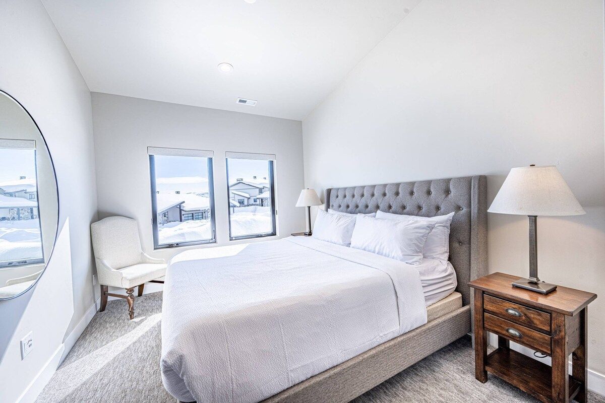 A modern bedroom with a large bed, a chair, a nightstand, and a window with a view.