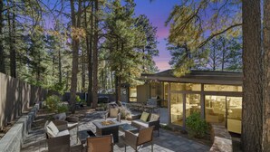 Une terrasse extérieure aménagée avec des meubles en osier, un foyer et un espace repas clos par des parois de verre, le tout entouré de grands arbres.