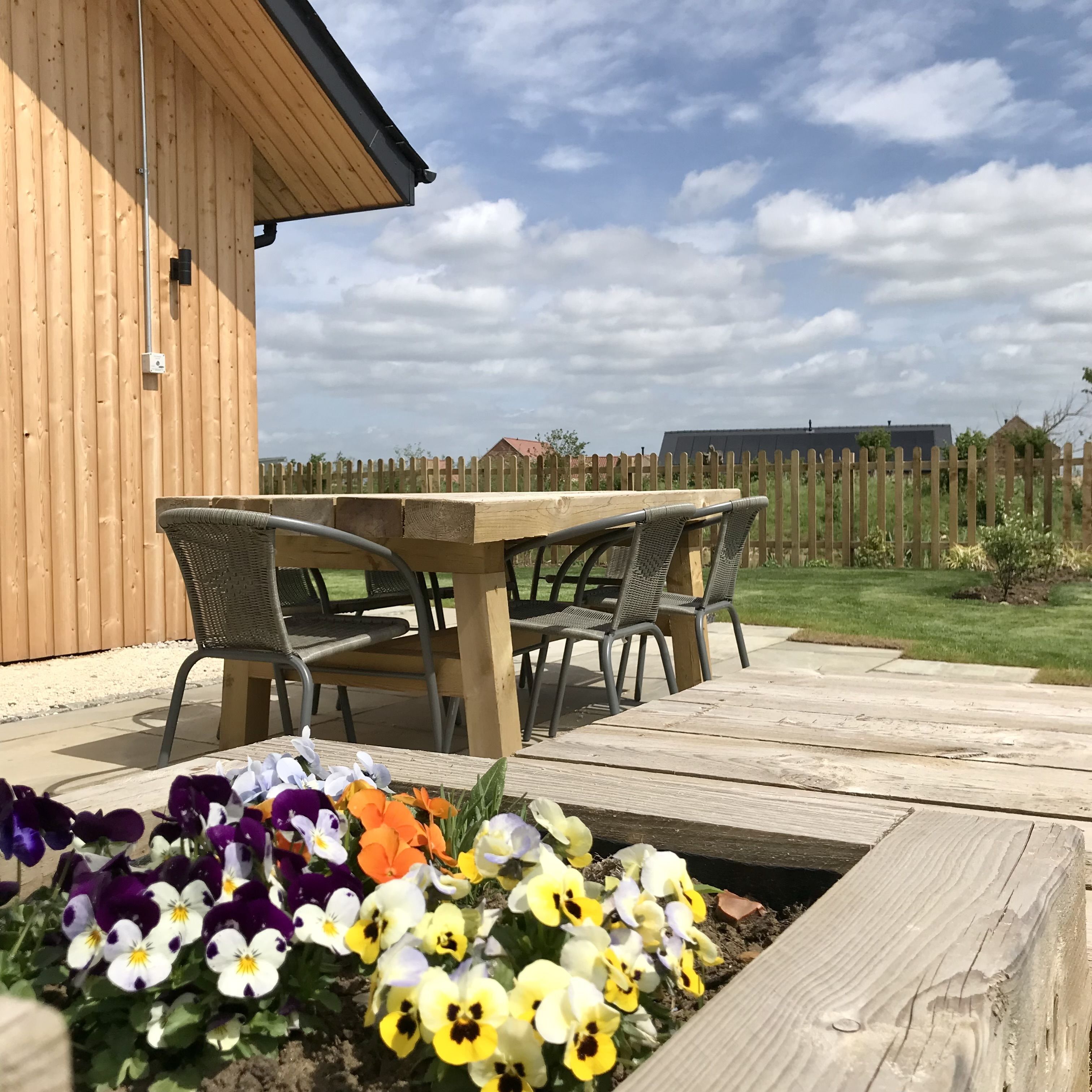 Eine Holzterrasse mit Tisch und Stühlen, umgeben von einem Garten mit Stiefmütterchen.