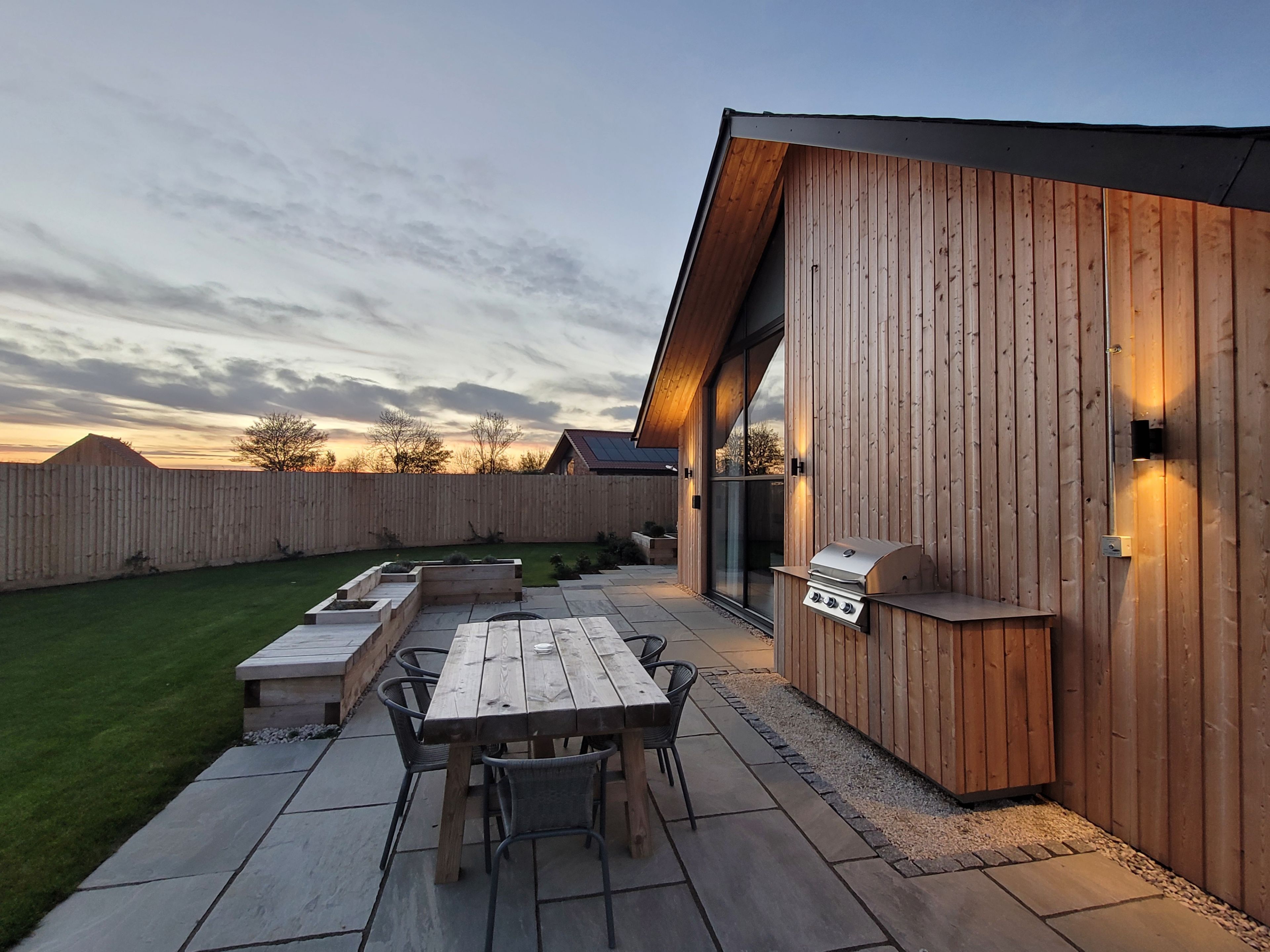 A patio with a wooden table and chairs, a barbecue grill, and a stone pathway.