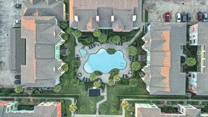 An aerial view of a residential area with a central swimming pool surrounded by houses, a parking lot, and a small garden area.
