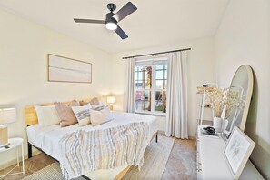 A bedroom with a bed, bedside table, lamp, ceiling fan, window with curtains, and a mirror.