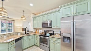 Una cocina con gabinetes verdes, electrodomésticos de acero inoxidable, encimeras de granito e iluminación colgante.
