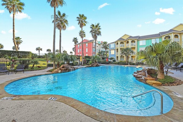 A swimming pool with a 5 ft depth marker, surrounded by palm trees and a multi-story building with balconies.