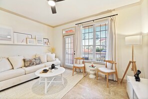 A modern living room with a sofa, coffee table, and two chairs. There is a window with blinds, a lamp, and framed pictures on the wall.