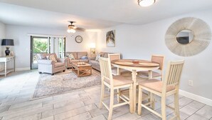 A modern living room with a sofa set, a coffee table, and a dining table with chairs. There is a ceiling fan, a wall-mounted mirror, and a window with a view of greenery. - South Tampa Retreat: Fenced Yard, Top Location! (Tampa)