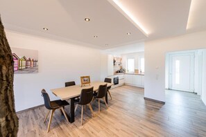 A spacious living area with a wooden floor, a dining table with chairs, a kitchen in the background, and a large window.