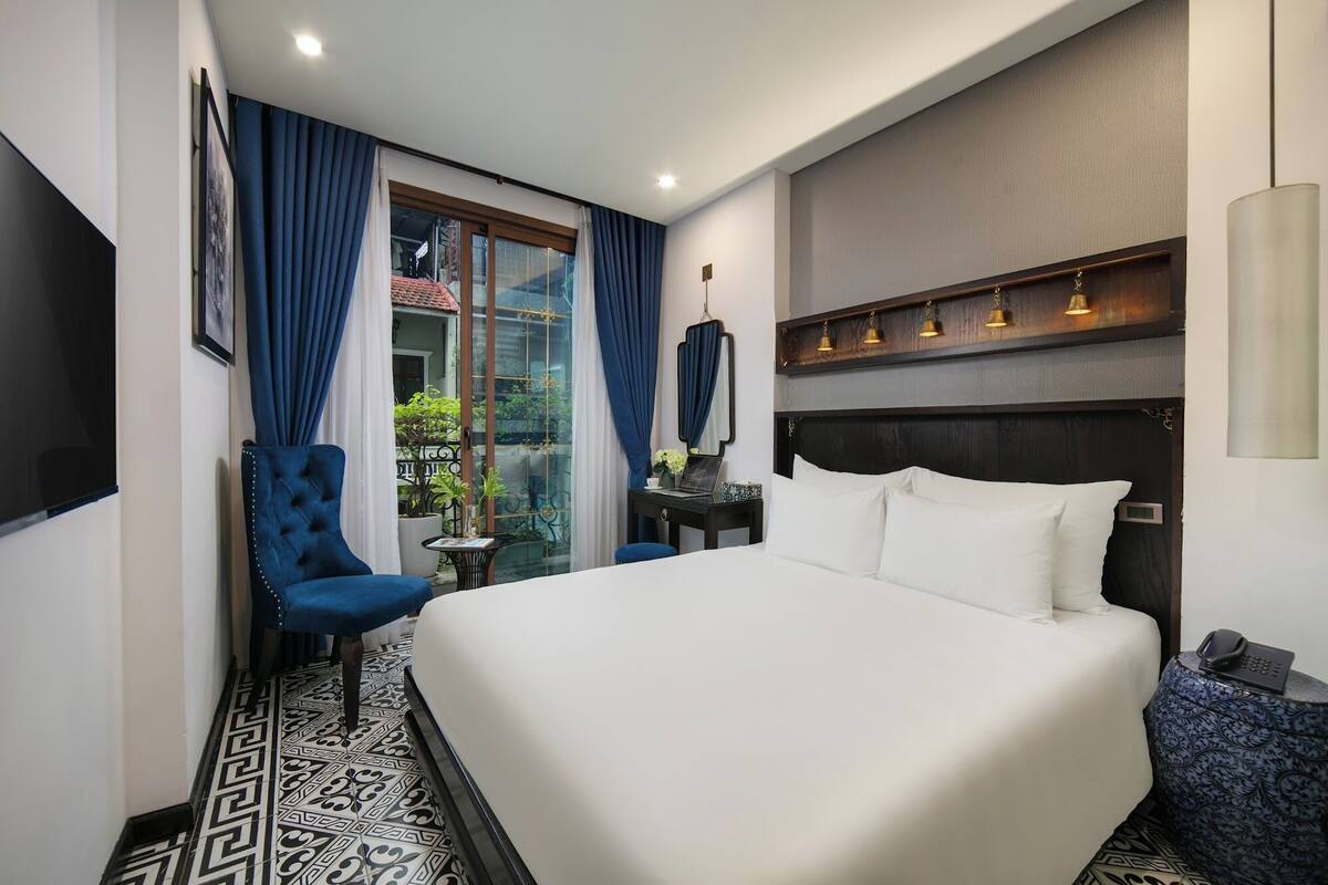 A hotel room with a large bed, a chair, a small table, and a window with curtains.