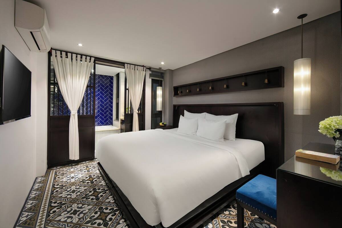 A modern hotel room with a large bed, a bedside table with a vase, and a patterned floor.